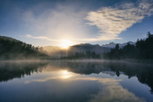 A calm mountain lake at sunrise with the reflection of the surrounding trees, mountains and sunrise showing on the lake.
