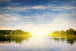Calm lake surrounded by lush trees with bright sunrise.