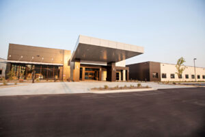 Cascadia of Nampa, front entrance. A concrete sidewalk and large drive-through portico attached to a long, one-story flat-roofed stucco building with many windows and flower beds between the sidewalk and the building.