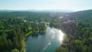 Aerial view of the Deschutes River in Bend, Oregon winding through the city surrounded by pine trees and grass under a hazy sky.
