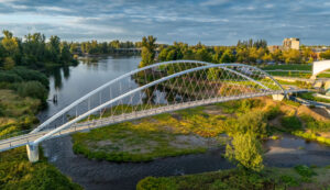 Aerial view of the Peter Minto Bridge crossing over the calm waters of the Willamette Slough in Salem Oregon on a partially cloudy day. Green grass and deciduous trees line the river with a view of the city in the background.