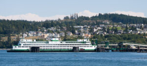 A large white and green ferry is docked at a terminal with a town on a tree-covered hill in the background.
