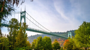 The steel-frame suspension St. John's Bridge against a lightly clouded sky with deciduous trees starting to show fall color beneath the bridge and in the background.