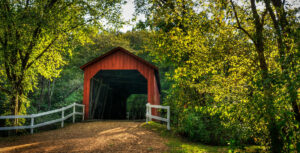 A dirt road leading to a red, wood bridge covered bridge surrounded by lush green deciduous trees.