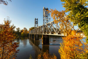 A narrow steel bridge that crosses a wide river that is lined with deciduous trees with leaves changing to fall colors.