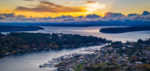 An early morning aerial view of the calm waters of Puget Sound with the sun rising above the cloud-covered mountains. Boat docks, houses and pine trees line the shores.