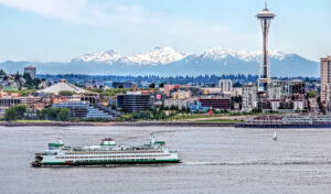 A ferry boat makes its way across the waters of the Puget Sound with city buildings and the Seattle Space Needle are in the background.