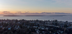 A waterfront city of houses among pine trees with a large body of water and a mountain range in the background at sunrise.